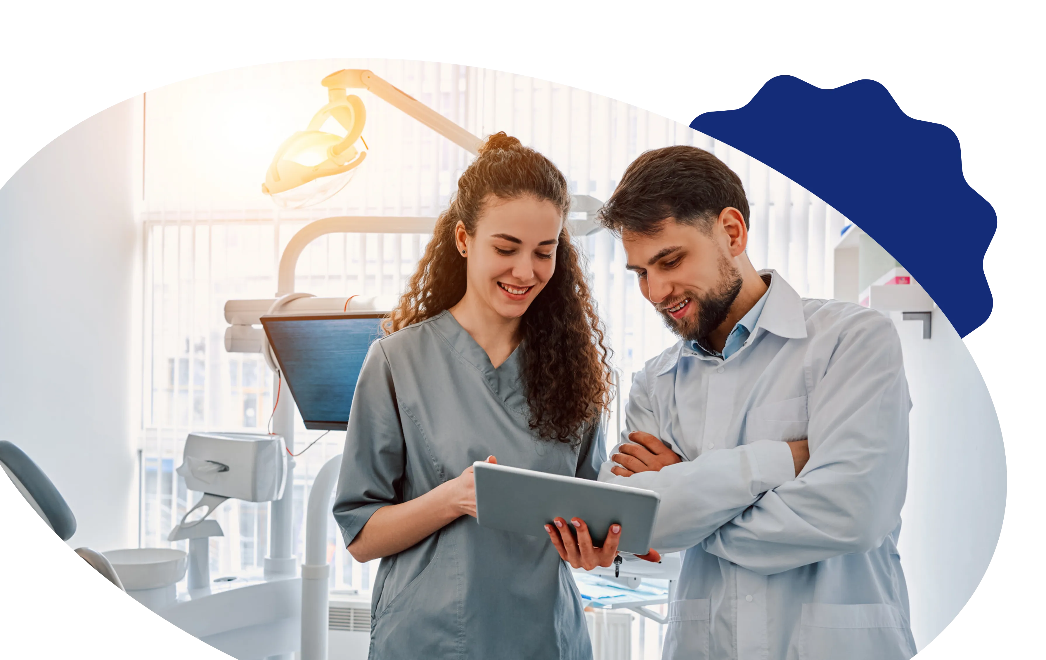 A dental professional and a hygienist review information on a tablet while standing in a modern dental office, framed within an oval-shaped design with a decoartive blue accent.